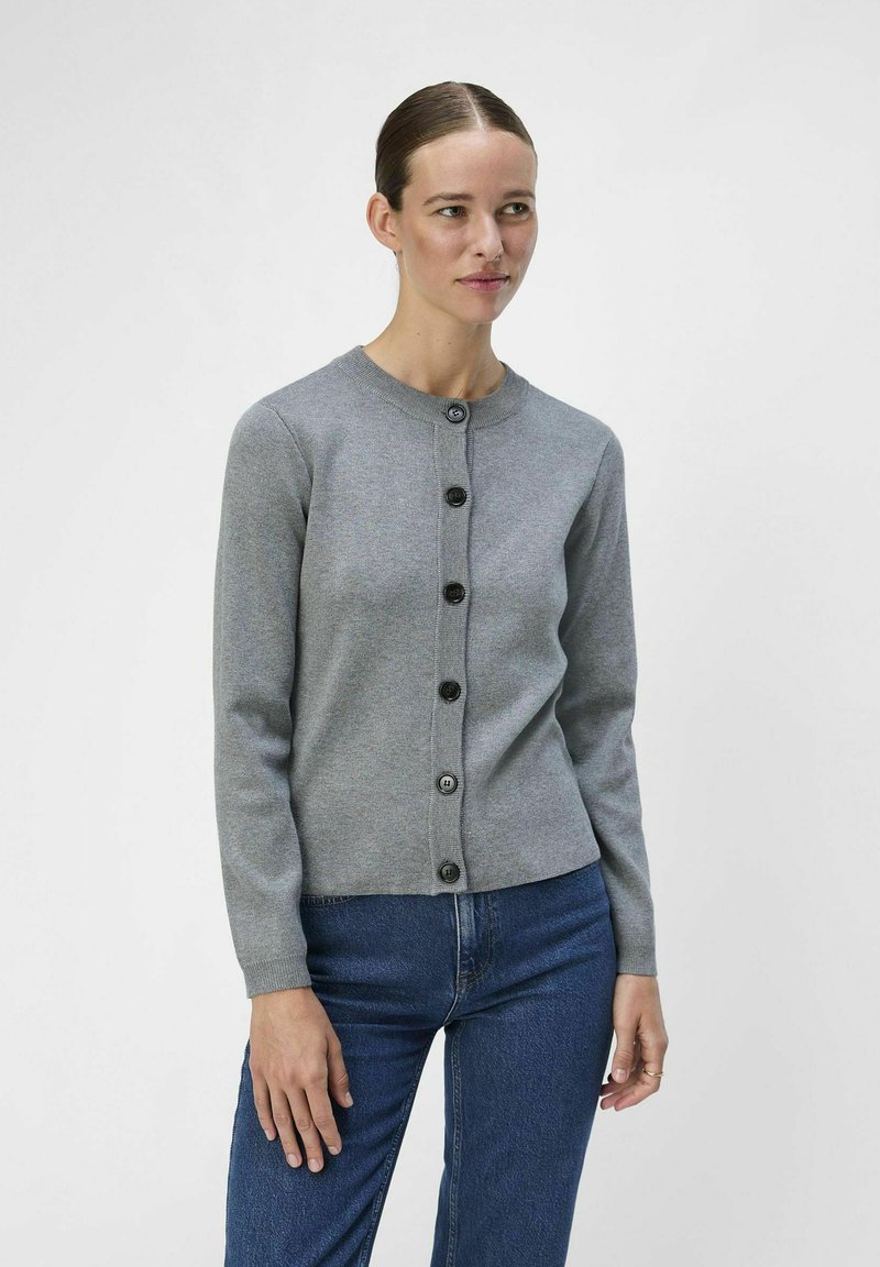 Grå strikket cardigan med rund hals, som har svarte knapper, lange ermer og en tettsittende design, sammen med blå jeans.