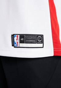 Maillot de basketball blanc avec accents rouges, présentant une étiquette noire indiquant la taille 48, le logo NBA et la marque Nike, avec une texture en maille.