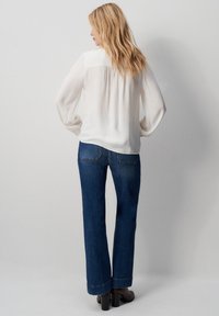 Blusa bianca fluida con maniche lunghe, abbinata a jeans blu scuro a gamba dritta. L'outfit presenta una vestibilità comoda e un design minimalista.