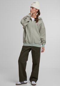 Jasnozielona bluza z zamkiem błyskawicznym i dużą kieszenią typu kangur, z białymi akcentami przeszyć, połączona z ciemnozielonymi spodniami oraz czarno-białymi trampkami.