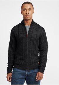Cardigan nero lavorato a maglia con zip frontale, caratterizzato da blocchi testurizzati sulla parte superiore del petto e polsini a coste, abbinato a jeans in denim blu.