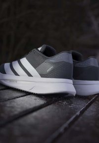 Ζευγάρι γκρι και λευκών παπουτσιών Adidas Adizero για τρέξιμο τοποθετημένα σε σκοτεινή ξύλινη επιφάνεια με θολό φόντο.