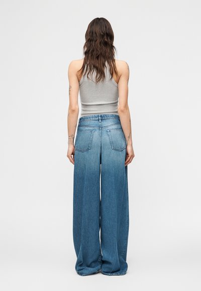 Γυναίκα με μακριά κυματιστά μαλλιά που φοράει γκρι αμάνικη μπλούζα και τζιν παντελόνι ψηλοκάβαλο με φαρδιά μπατζάκια, όρθια με την πλάτη γυρισμένη σε λευκό απλό φόντο.