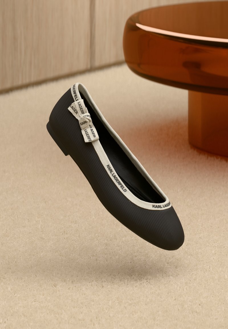 Sapato plano preto com textura canelada, acabamento creme e um laço decorativo. Logótipo visível no acabamento. Colocado sobre uma superfície clara e felpuda.