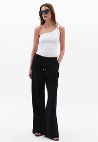 Top blanco de un solo hombro con detalle de nudo, combinado con pantalones de pierna ancha negros que cuentan con una cintura ajustable y dos cuentas plateadas como acento.