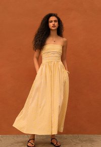 Donna con capelli ricci che indossa un vestito giallo e bianco a quadretti senza spalline, sandali neri, in piedi contro un muro arancione con texture.