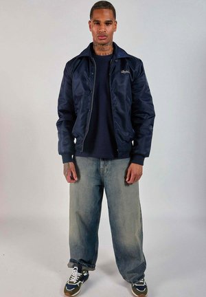 Homme debout, face à l’avant, portant une veste bleu marine, une chemise noire, un jean ample délavé et des baskets noires et blanches sur un fond uni.