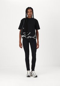 Crna oversized majica s bijelim "Karl" logotipom, uparena s crnim skinny trapericama i tenisicama s srebrnim detaljima. Ima pletenu kosu i torbu preko ramena.