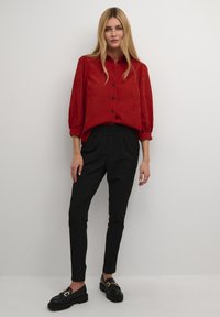 Blouse rouge à boutons avec des manches bouffantes ; associée à un pantalon noir ajusté. Mocassins noirs avec des accents en or. Fond uni.