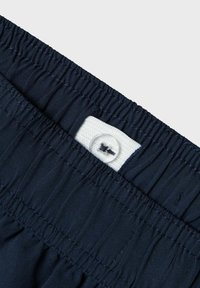 Granatowa elastyczna talia z marszczonym teksturą, z białą metką z tkaniny i przezroczystym guzikiem. Gładki, lekki materiał.