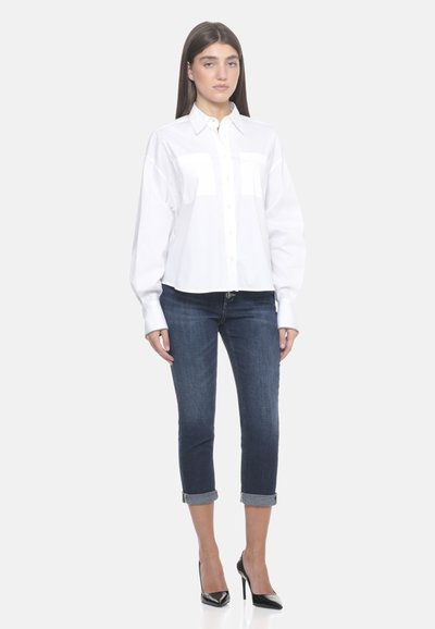 Camicia bianca con bottoni, maniche lunghe e due tasche sul petto, abbinata a jeans cropped scuri e scarpe con tacco nere.