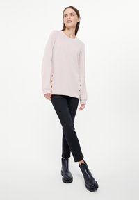 Lichtroze langemouwen top met gouden knopen aan de zijkant, gecombineerd met zwarte slim-fit broek en zwarte enkellaarsjes. Eenvoudig, modern ontwerp.