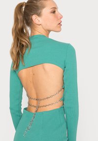 Top turquesa canelado com mangas longas, apresentando costas abertas e detalhes de corrente prateada na cintura. Textura suave, design ajustado.