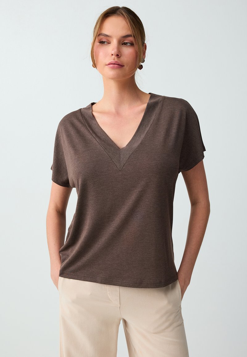 Femme portant un haut marron à col en V manches courtes et un pantalon beige, debout, mains dans les poches, sur un fond clair uni.