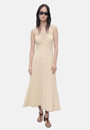 Cream-colored, ankle-length dress with spaghetti straps and intricate texture. Features a subtle pattern and delicate lace trim at the hem.