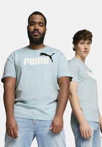 Camisetas de algodón azul claro con la marca "PUMA" en blanco y el logo del puma en negro. Mangas cortas, cuello redondo, textura suave, combinadas con jeans de mezclilla claros.