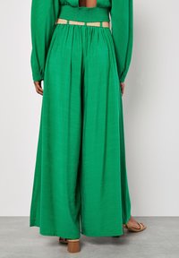 Groene wijde broek van gladde stof, met een geplooide tailleband, een beige riem en een vloeiende silhouet tot aan de enkels.