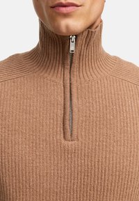 Brun stickad tröja med hög krage, ruggad struktur och halvdragkedja framtill. Tyget verkar mjukt och strukturerat.