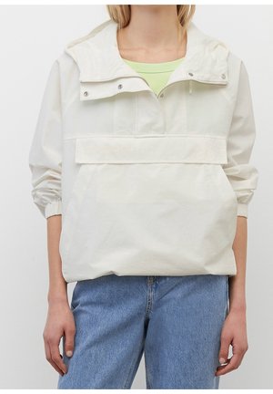 Personne portant un coupe-vent blanc à capuche avec une poche avant à rabat et une chemise vert clair en dessous, associé à un jean bleu.