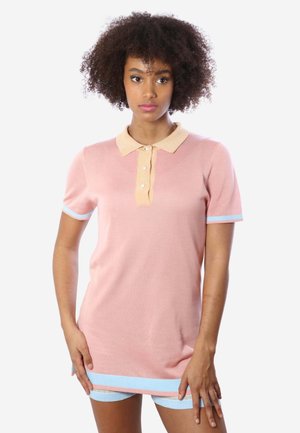 Giovane donna con capelli ricci che indossa una polo rosa con colletto beige e bordini blu, in piedi davanti a uno sfondo bianco semplice.