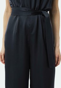 Zwarte jumpsuit met een brede, gebelte taille, geplooide voorkant en vloeiende stof. Glad textuur met een strakke, moderne uitstraling.