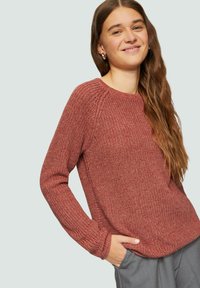 Roter strukturierter Strickpullover mit Rundhalsausschnitt, langen Ärmeln, gerippten Bündchen und lockerer Passform. Das Model hat die Hände in den Taschen einer grauen Hose.