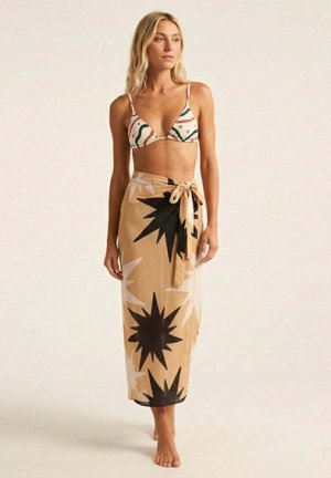 Mujer de pie descalza, vistiendo un top de bikini con estampado y una falda cruzada beige con grandes diseños de estrellas negras y blancas.