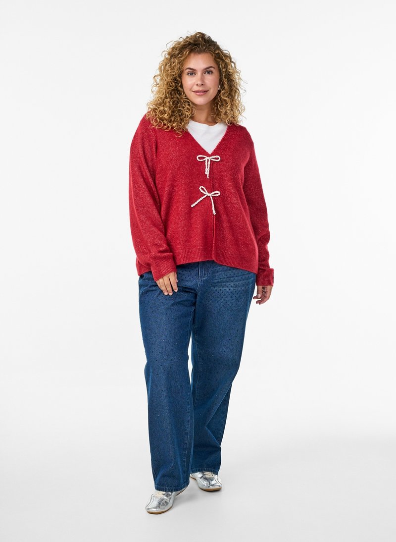 Frau mit lockigem Haar, die einen roten Cardigan mit weißen Bändern, ein weißes Hemd, blaue Jeans und silberne Schuhe trägt, steht vor einem weißen Hintergrund.