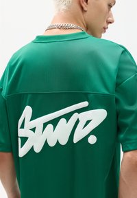 Tricou sportiv din plasă verde cu mâneci scurte, având un logo alb proeminent pe spate și o textură netedă, respirabilă.