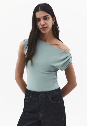 Ung kvinde med langt mørkt hår iført en lysegrøn top med én skulder og sort højtaljet jeans, stående med hænderne på ryggen.