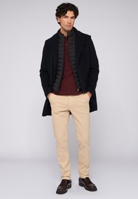 Schwarzer Mantel, darunter eine gesteppte schwarze Weste, burgunderfarbenes Polohemd, khakifarbene Hose und dunkelbraune Slipper. Glatte Stoffe mit strukturiertem Design.