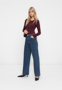 Donna con lunghi capelli biondi, che indossa un top fitted a maniche lunghe color bordeaux, jeans blu a vita alta e a gamba larga, tacchi neri a punta e una piccola borsa a tracolla nera.