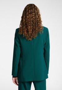 Blazer verde con una vestibilità su misura, spalle strutturate, tessuto testurizzato e una singola apertura sul retro. Abbinato a pantaloni coordinati.