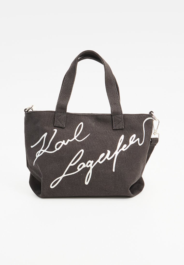 Bolso tote de lona negra con asas cortas, que presenta el texto en cursiva blanca "Karl Lagerfeld" en la parte frontal. Cierre con cremallera y correa desmontable.