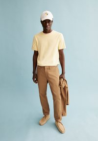 Helles gelbes T-Shirt mit kurzen Ärmeln, beige Chinos und beigefarbene Mokassins. Weiße Mütze mit blauem Design. Einfarbiger blauer Hintergrund.