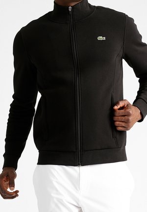 Schwarze Reißverschlussjacke mit grünem Krokodil-Logo, getragen von einer Person mit dunkler Haut und weißer Hose, Hand nahe der Tasche auf der rechten Seite platziert.