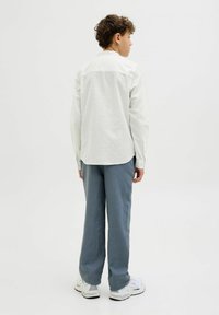Camicia bianca a maniche lunghe con orlo arrotondato, indossata con pantaloni azzurri chiari e sneaker bianche; il tessuto sembra leggero e con texture.