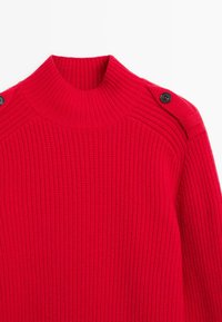 Světle červený pletený svetr s vysokým límcem a černými knoflíky na ramenních popruzích, položený na bílém pozadí.