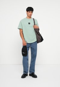 T-shirt kratkih rukava u mint zelenoj boji s crnom aplikacijom loga, uparen s svijetloplavim trapericama, crnom kapom u ruci i tamnim cipelama.