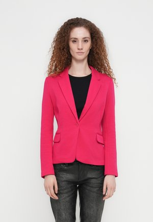 Ženska z kodrastimi lasmi nosi svetlo rožnato suknjič čez črno majico in temne teksturirane hlače, stoji na enobarvni svetli podlagi.