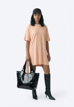 Vrouw draagt een oversized perzikkleurige t-shirtjurk, zwarte pet, zwarte kniehoge laarzen en houdt een grote glanzende zwarte totebag vast.