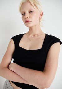 Černý top s krátkým rukávem a čtvercovým výstřihem. Modelka má na sobě vrstvené zlaté a perlové náhrdelníky na jednobarevném bílém pozadí.