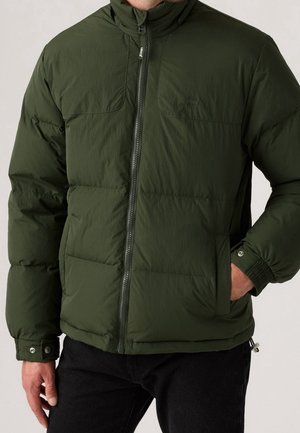 Homme portant une doudoune vert foncé avec une fermeture éclair à l'avant et des poignets à boutons-pression, une main dans la poche de la veste.