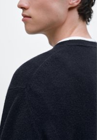 Tmavě námořnický svetr, jemná textura, kulatý výstřih, žebrované manžety, s minimalistickým designem a mírně volným střihem. Pohled zezadu a na rameno.