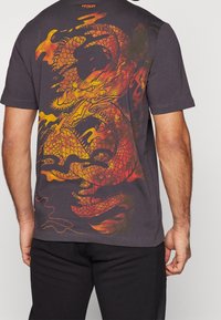 Camiseta gris de manga corta con un diseño colorido de dragón en naranja y amarillo en la parte posterior. Hecha de una tela suave con un ajuste cómodo.