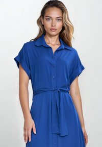 Niebieska sukienka koszulowa z gładkiego materiału, z zapięciem na guziki z przodu, krótkimi podwiniętymi rękawami oraz dopasowanym paskiem w talii.