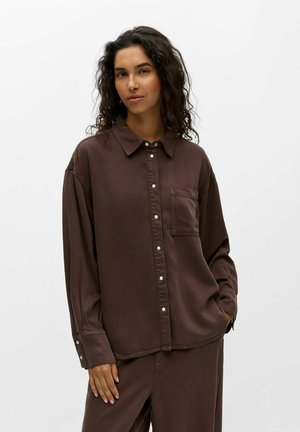 Femme aux cheveux bouclés et foncés portant une chemise marron ample boutonnée avec une poche poitrine et un pantalon marron assorti sur un fond clair uni.