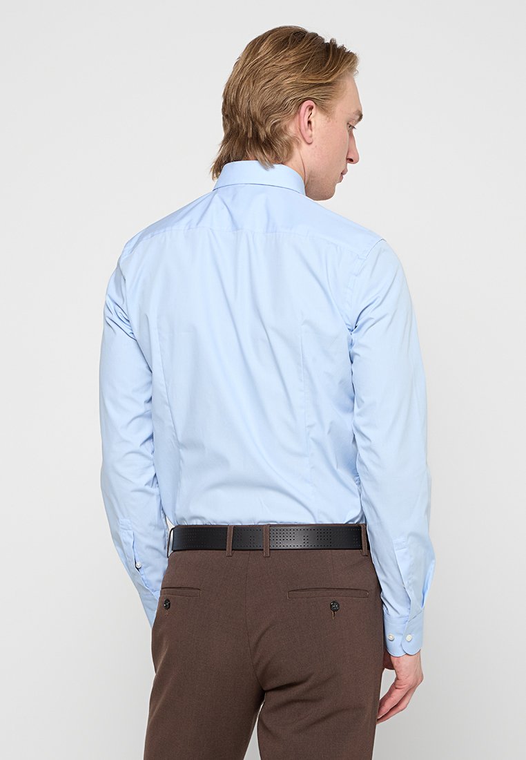 Camicia azzurra chiara con colletto classico, maniche lunghe e pieghe posteriori, abbinata a pantaloni marroni e una cintura perforata nera.