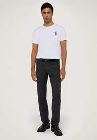 Bílá tričko s černým grafickým potiskem, spárované s tmavě šedými kalhotami. Černý opasek a lesklé tmavé boty dotvářejí outfit.