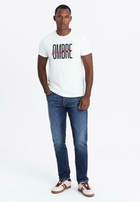 Camiseta blanca de manga corta con la palabra "OMBRE" impresa en letras negras y rojas, combinada con jeans azules y zapatillas blancas con detalles en marrón.